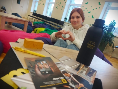 Daugavpils Tehnoloģiju un tūrisma tehnikuma izglītojamā piedalās SIA „MSC Shared Service Center Riga” tiešsaistes lekcijā par projektu vadību, fonā redzami uzņēmuma informatīvie materiāli un dāvanas.