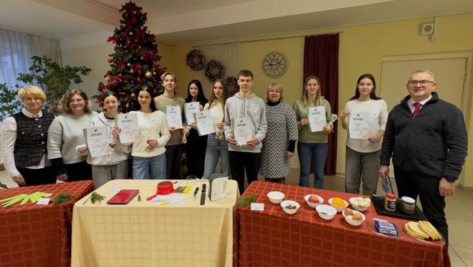 Grupas foto ar konkursa dalībniekiem, skolotājiem un komisijas locekļiem, izglītojamie tur diplomus un balvas, priekšplānā redzami konkursa uzdevumu galdi.