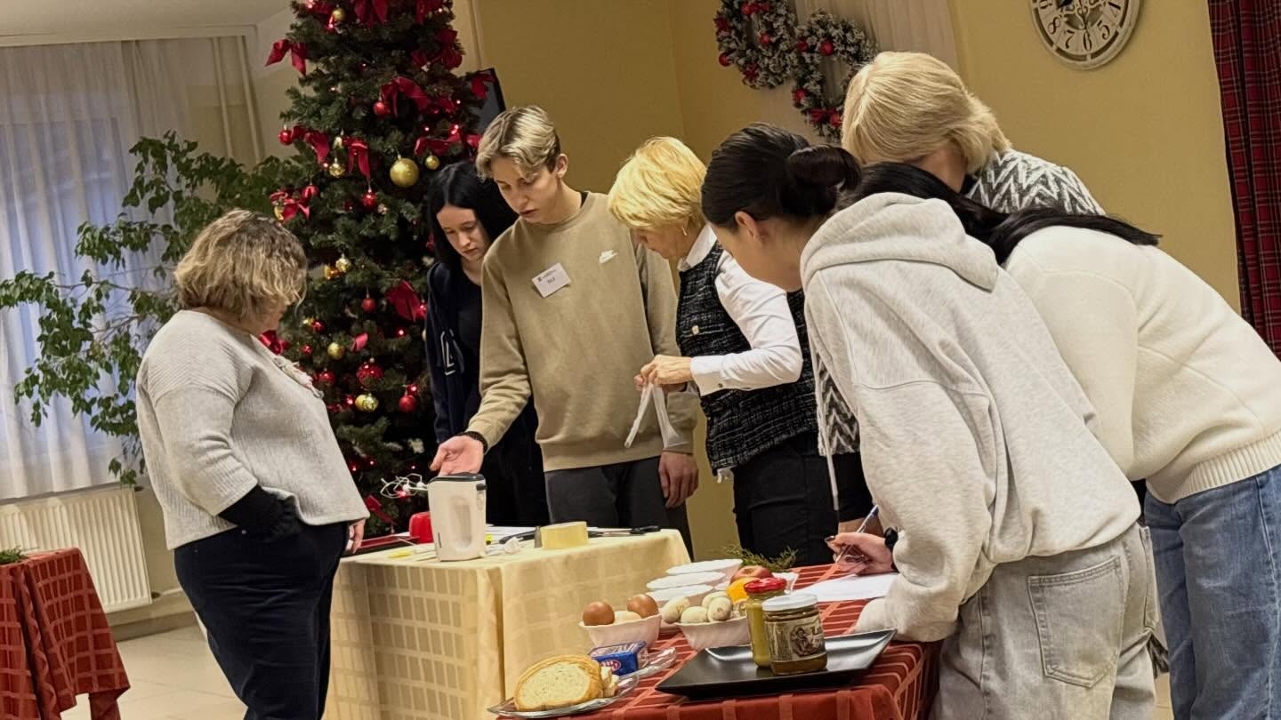 Konkursa dalībnieki un skolotāji pulcējušies pie galda ar inventāru un produktiem, apspriežot uzdevumu izpildi Ziemassvētku noformētā telpā.