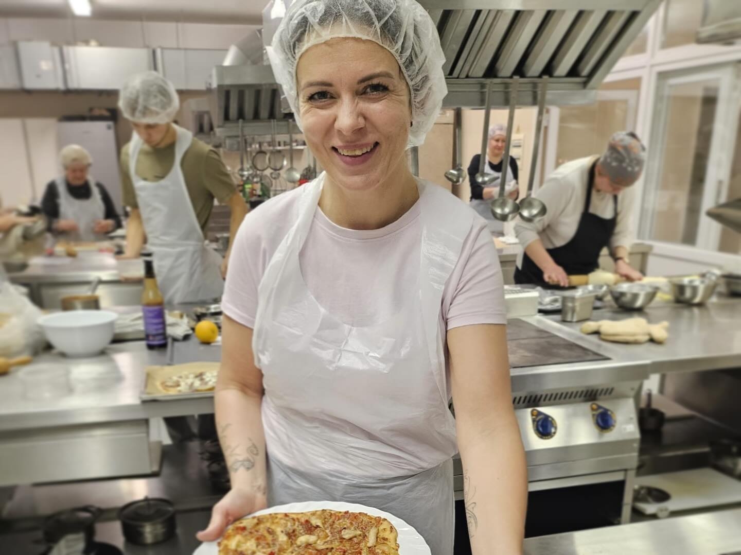 Neformālās izglītības kursa vadītāja pasniedz dalībniekiem svaigi ceptu picu, daloties ar profesionāliem padomiem ēdienu gatavošanā.