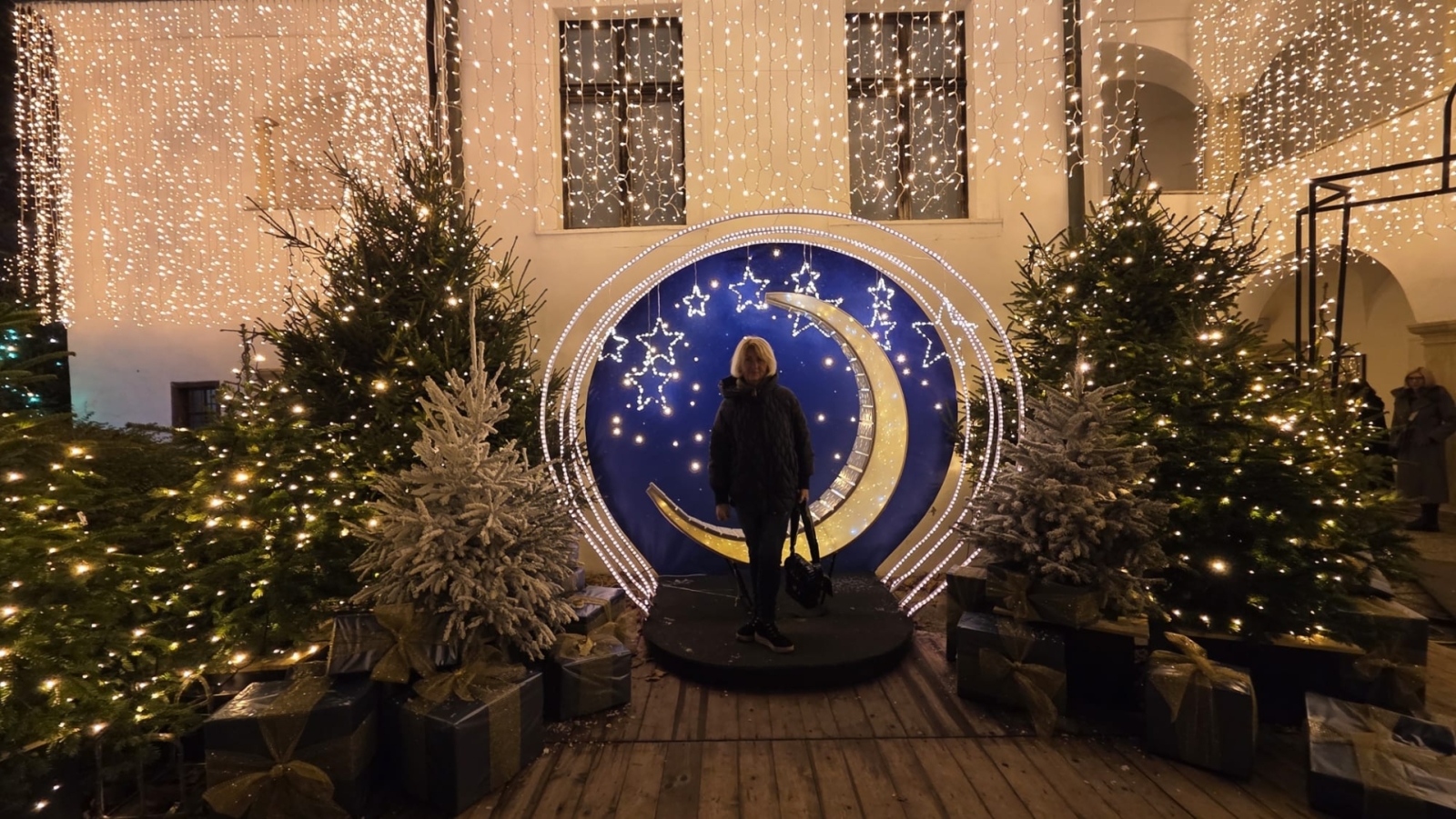 Projekta dalībniece pie Ziemassvētku foto zonas ar dekoratīvu mēnesi un izgaismotām eglēm.