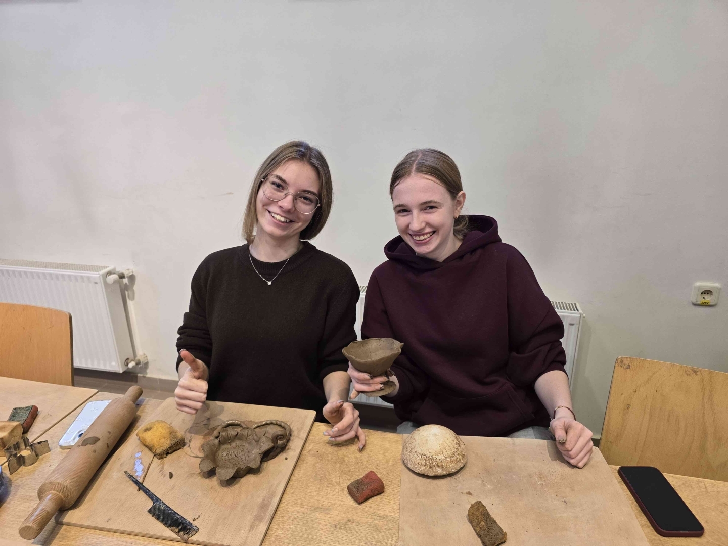 Divas izglītojamās smaida un demonstrē savus no māla veidotos darbus keramikas nodarbībā