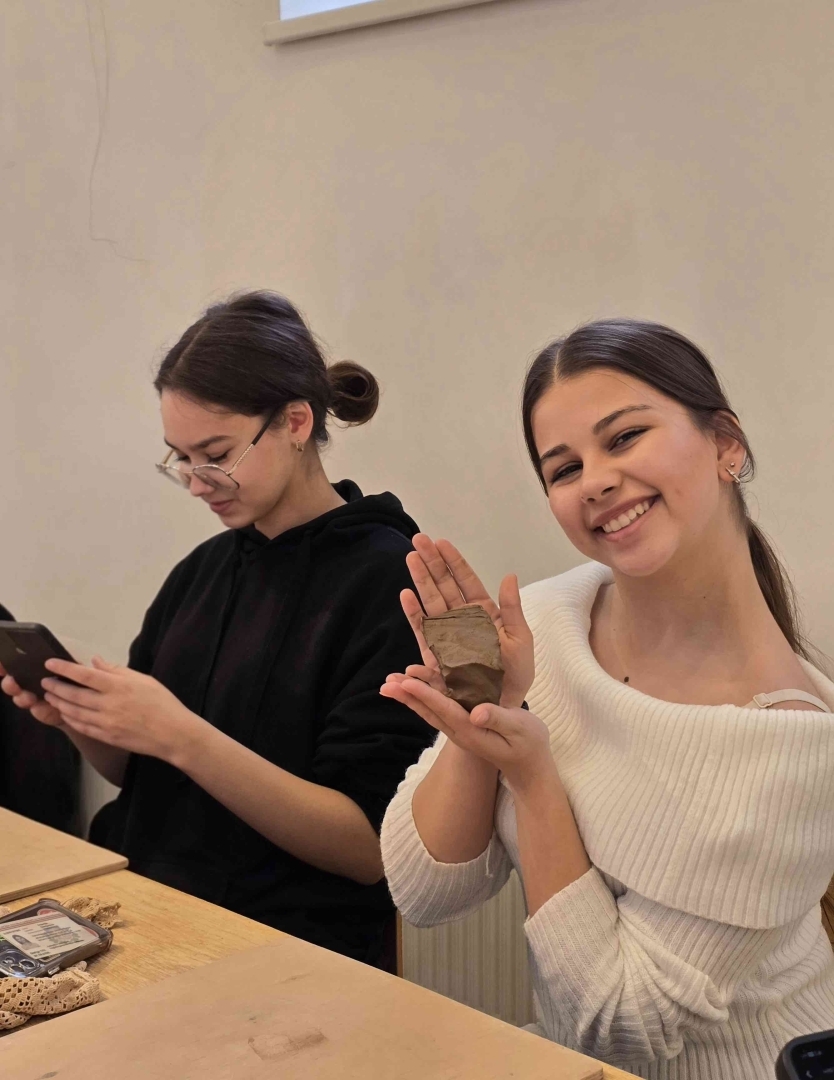Izglītojamā rāda rokās turētu māla izstrādājumu, kamēr cita dalībniece strādā ar telefonu