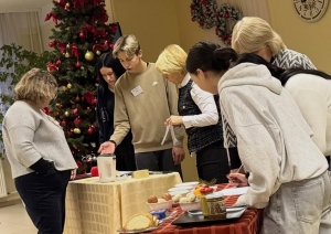 Konkursa dalībnieki un skolotāji pulcējušies pie galda ar inventāru un produktiem, apspriežot uzdevumu izpildi Ziemassvētku noformētā telpā.